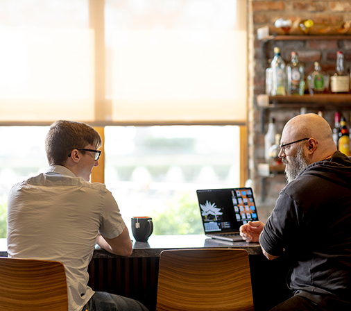 Two people sitting at a sunlit bar with a coffee cup and laptop near them